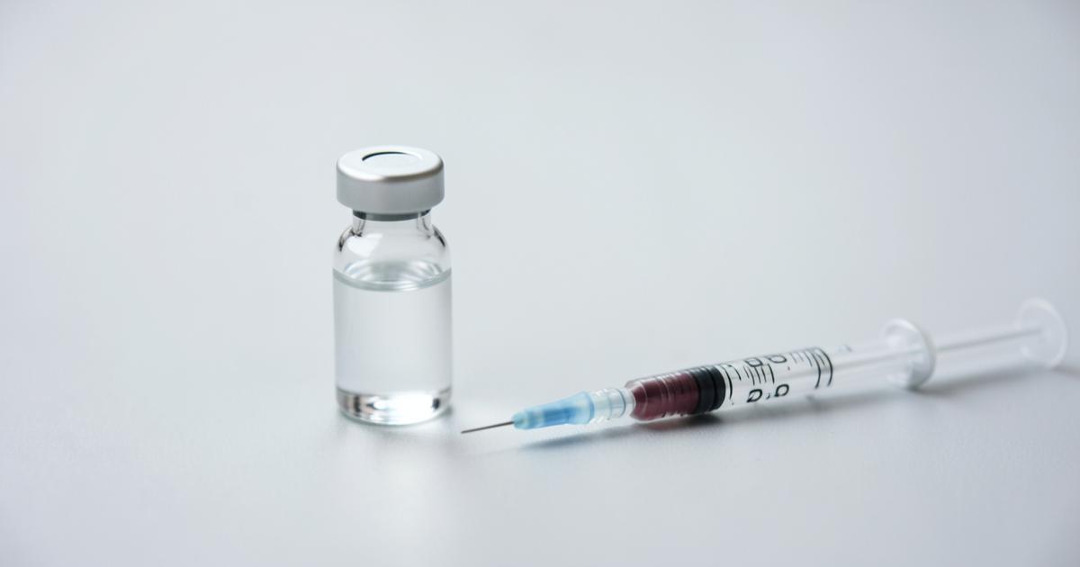 A clear glass vial of solution beside an insulin syringe on a pale surface