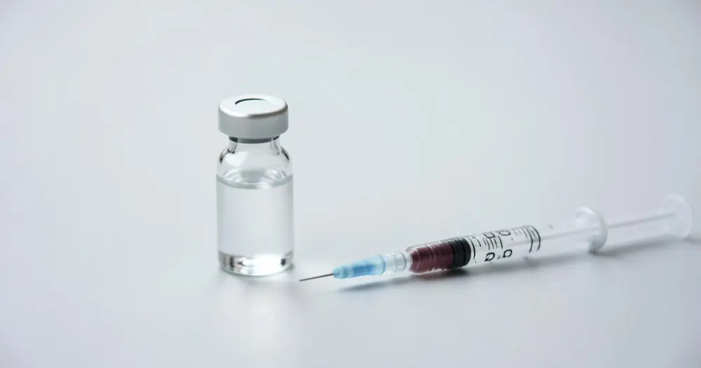 A clear glass vial of solution beside an insulin syringe on a pale surface A clear glass vial of solution beside an insulin syringe on a pale surface