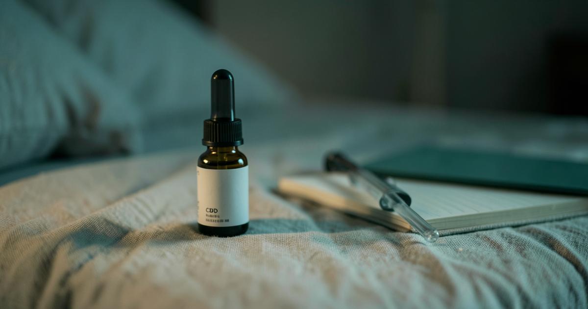 A dropper bottle of CBD oil with a glass pipette on a linen nightstand at night