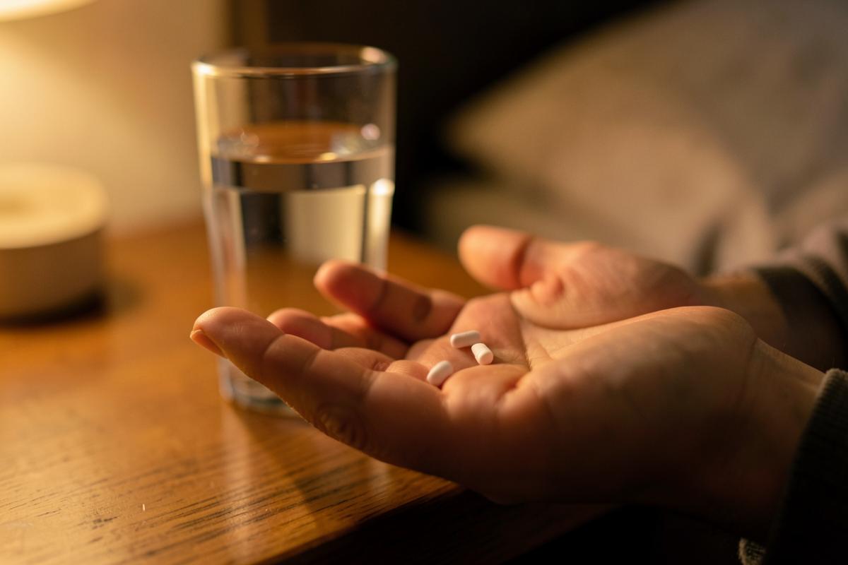 Two small tablets in an open palm beside a glass of water on a nightstand