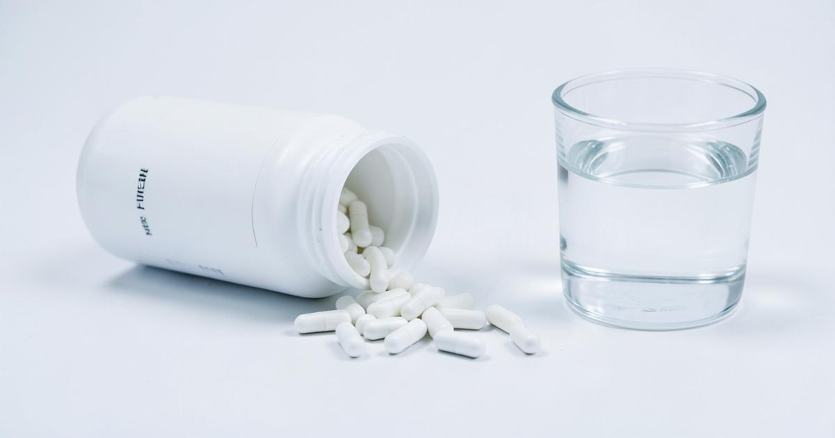 Plain white capsules spilling from an unlabelled bottle beside a glass of water